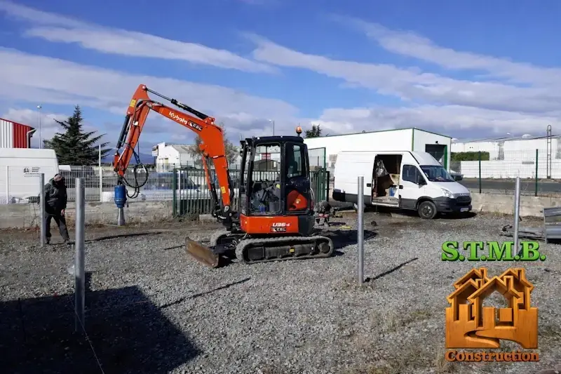 Chantier de construction avec mini pelle installant des poteaux metalliques sur terrain gravillonne