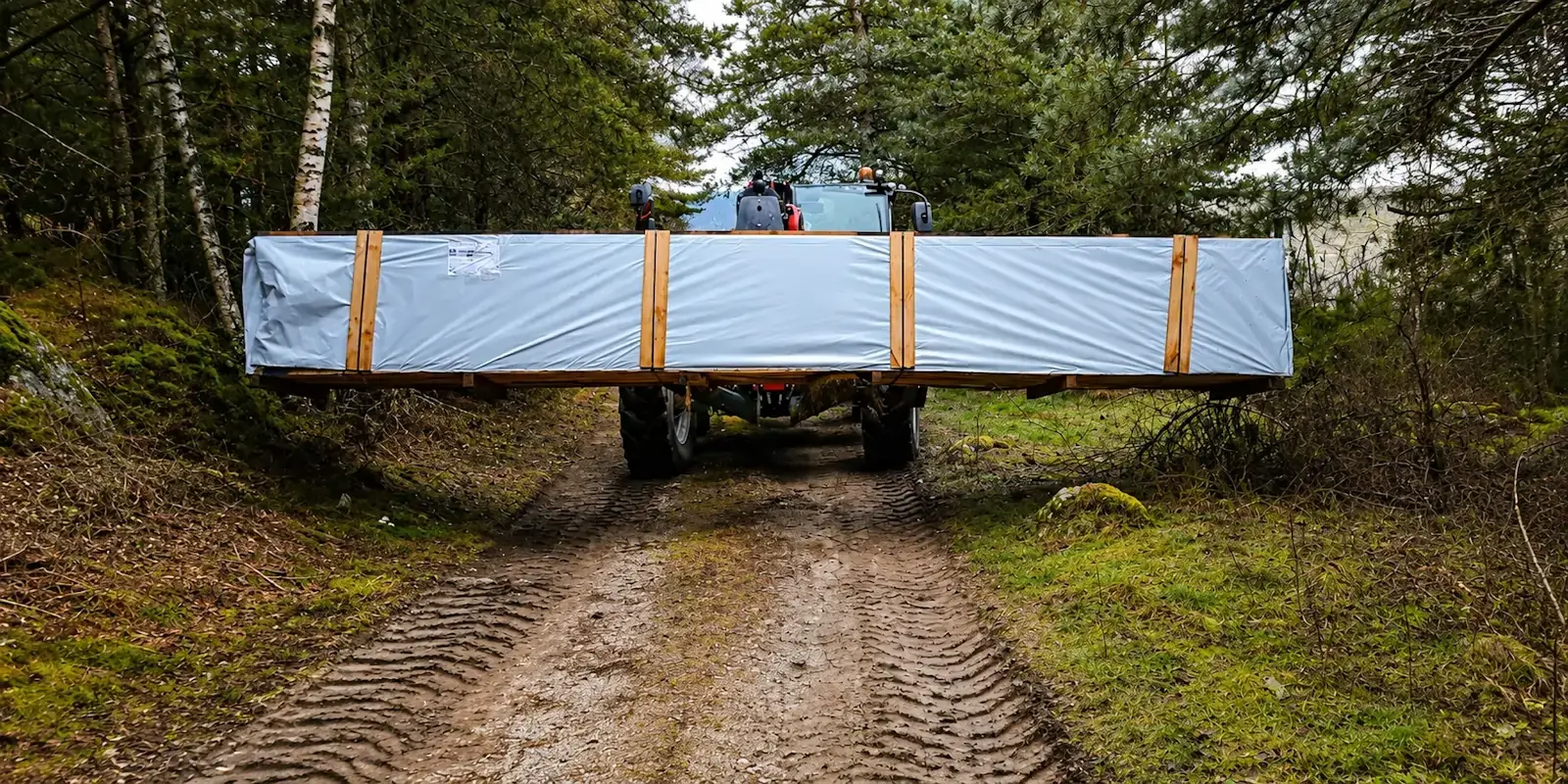 Livraison d'un chalet en bois sur un chemin forestier avec un chariot tout-terrain pour accéder à un terrain difficile.